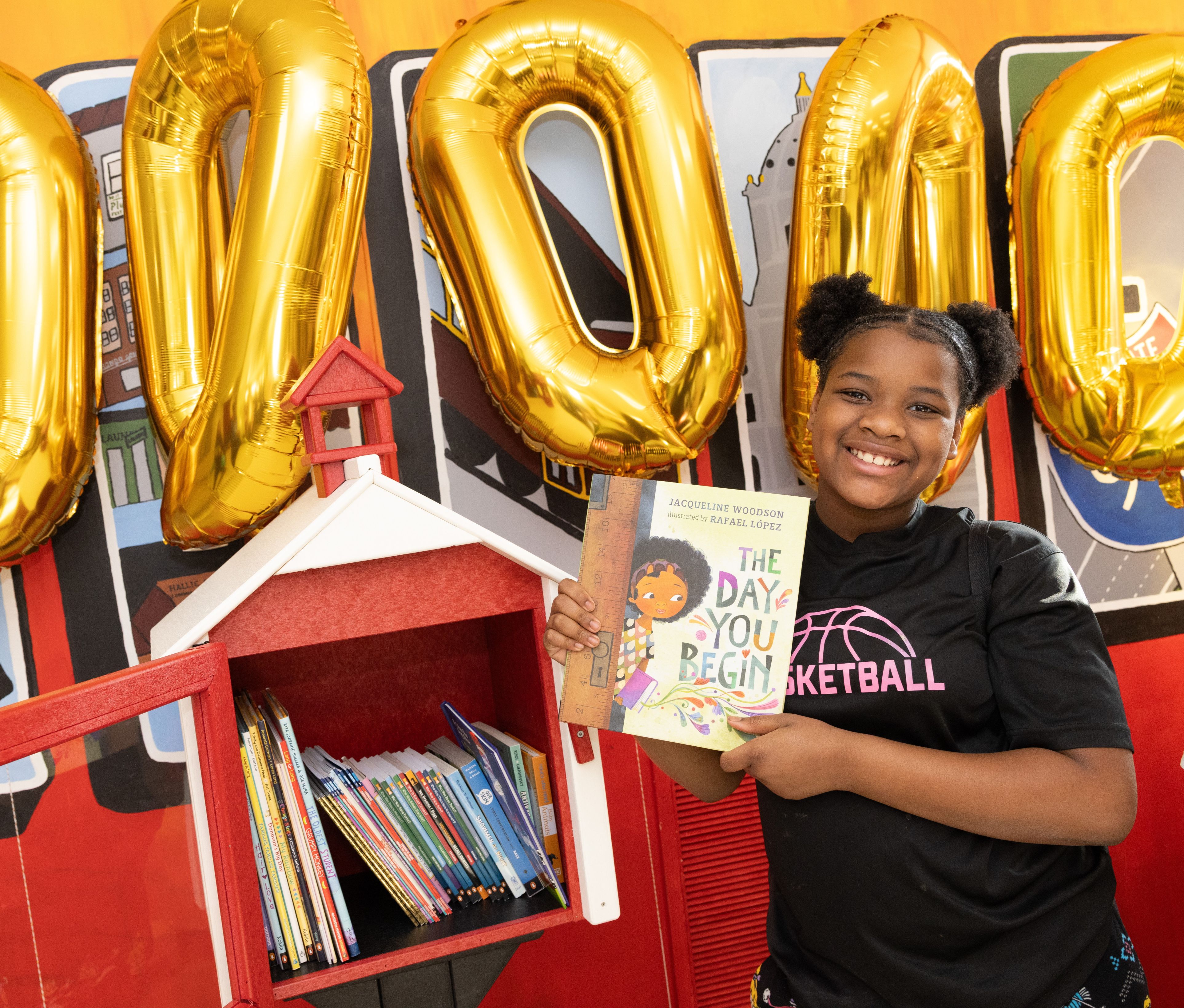 A student holds a book in front of the 200,000th Little Free Library book-sharing box.